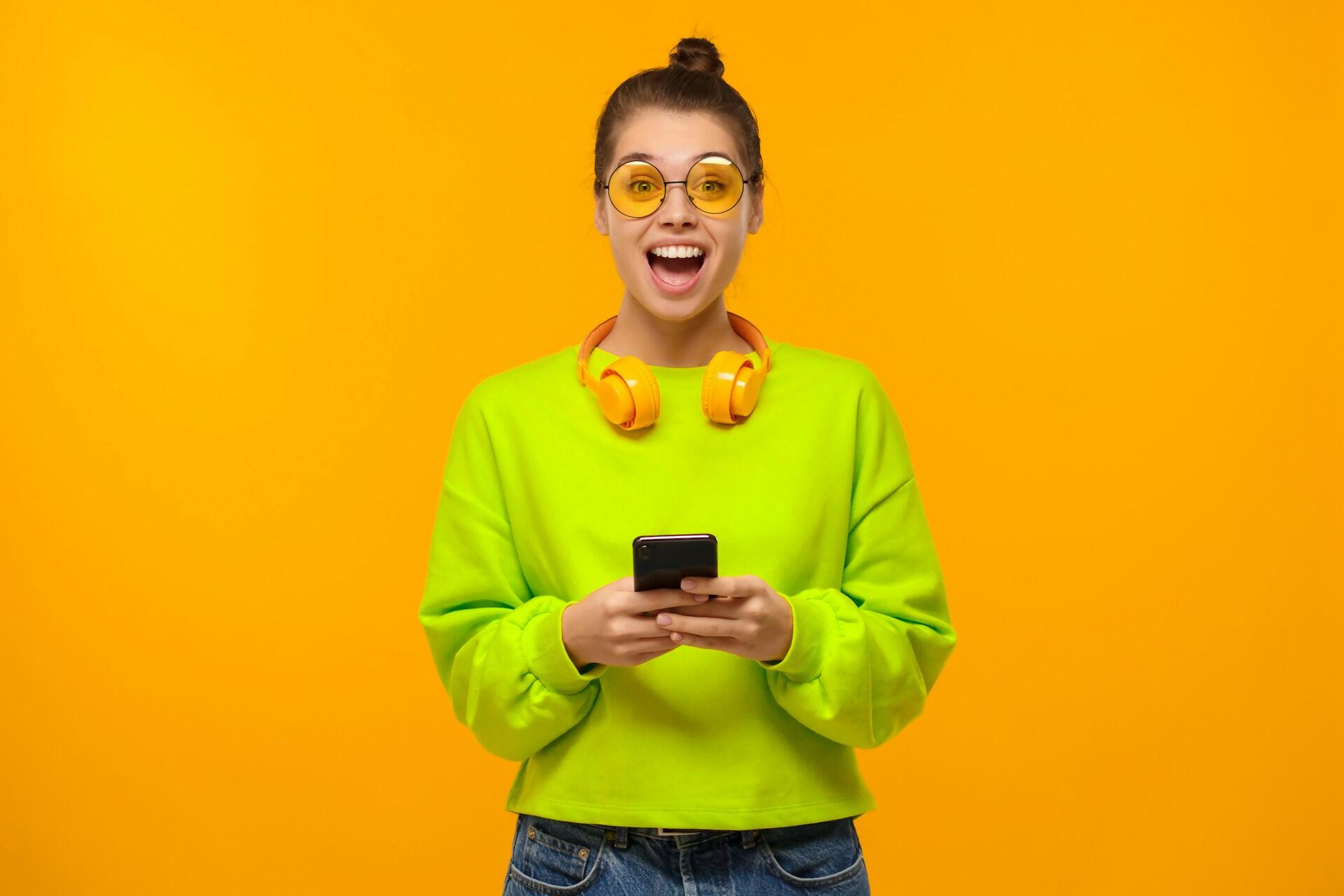 A person in a bright jumper holds a smartphone and looks happy and surprised.